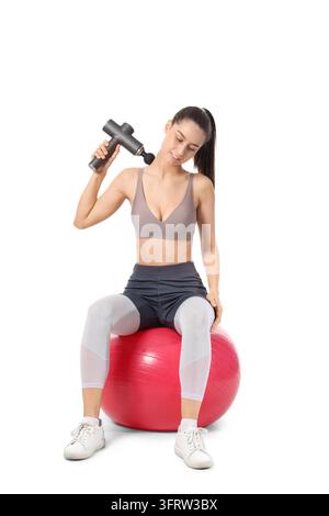 Muskelerholung und Physiotherapie, Frau verwendet eine Massagpistole am Nacken, während sie auf einem Fitnessball sitzt, zur Schmerzlinderung und Wohlbefinden in einer Studio-Umgebung Stockfoto