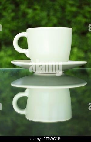 Symmetrische Reflexion einer weißen Keramikkaffeetasse auf einem Glastisch, Gleichgewichtsbegriff mit verschwommenem grünen Blattgrund Stockfoto