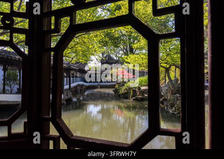 Klassischer Gartenteich mit einem weiß verkleideten Pavillon, gesehen durch ein traditionelles hölzernes Gitterfenster, Garten des bescheidenen Verwalters, Suzhou, China Stockfoto