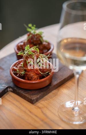 Gourmet-Spanische Fleischbällchen in reichhaltiger Tomatensoße, serviert in keramischen Terracottacazuelas, garniert mit Mikrogreens neben Weißwein. Stockfoto