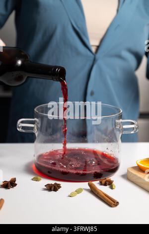Nahaufnahme in einer gemütlichen Heimküche zeigt den Vorbereitung von Glühwein: Rotwein wird in einen Glastopf mit Zimt, Anis, OrangenSchale und Kardamom gegossen, hi Stockfoto