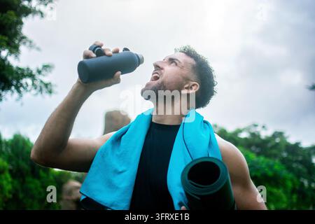 Niedrigwinkelaufnahme eines sportlichen Mannes, der Wasser aus einer wiederverwendbaren Sportflasche trinkt, während er eine gerollte Yogamatte trägt und ein blaues Handtuch um den Nacken trägt Stockfoto