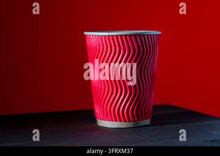 Roter wellenförmiger Pappkaffeebecher auf dunklem Holztisch. Nahaufnahme eines roten, gerippten Einweg-Pappbechers für Heißgetränke. Zum Mitnehmen-Kaffeebecher auf dunklem Holz de Stockfoto