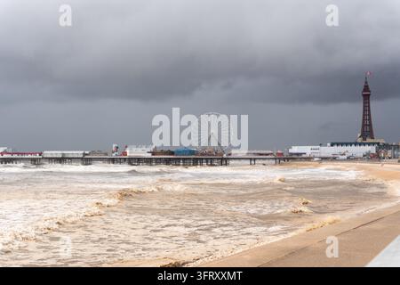 Dramatische raue braune Wellen, die gegen die Promenade schlagen, mit dem Central Pier, dem Riesenrad und dem Blackpool Tower unter dunklen Sturmwolken. Stockfoto