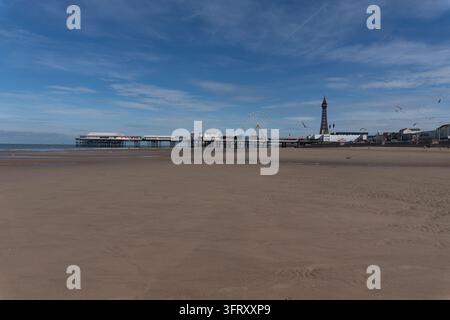 Ein weitläufiger Blick über einen leeren goldenen Sandstrand in Richtung des Central Pier-Riesenrads und des Blackpool-Turms unter einem klaren blauen Himmel. Stockfoto