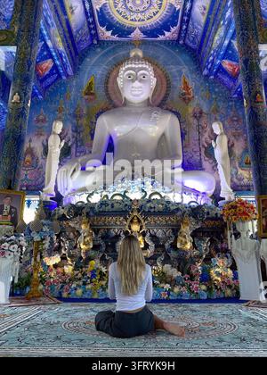 Inneres des Blauen Tempels in Chiang Rai, Thailand, mit einer jungen blonden Touristin, die vor der imposanten weißen Haupt-Buddha-Statue meditiert. Stockfoto