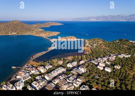 Luftaufnahme eines Küstendorfes und einer Halbinsel nahe Elounda, Kreta, mit tiefblauem Meer und Salinen, die einen schmalen Damm säumen, weitläufige Landschaftsaufnahme Stockfoto