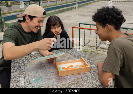 Mann mit seiner Nichte und einem Freund, die an einem Parktisch sitzen und ein Brettspiel spielen, während sie gemeinsam Zeit im Freien verbringen in einer fröhlichen und lustigen Familienszene Stockfoto