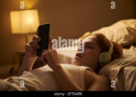Mann mit Kopfhörern, der ein Smartphone benutzt, während er im Bett liegt Stockfoto