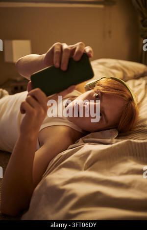 Junger Mann liegt auf dem Bett und verwendet ein Smartphone mit Kopfhörern Stockfoto