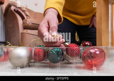 Nahaufnahme von reifen Händen in einem gelben Pullover, die rote, türkisfarbene und silberne Kugeln aus einer unterteilten durchsichtigen Plastikaufbewahrungsbox auswählen, um zu dekorieren Stockfoto