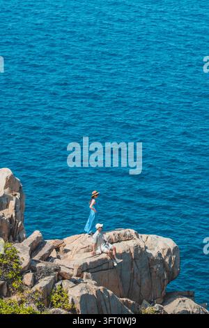 Paar ruht auf Granitfelsen hoch über dem tiefblauen Meer, Frau in blauem Kleid und Strohhut, Con Dao, Vietnam Stockfoto