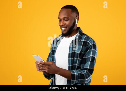 Musikanwendung. Afrikanischer Mann, Der Das Telefon Benutzt, Ohrstöpsel Trägt Und Lied Online Hört, Stehend Über Gelbem Studio-Hintergrund. Smartphone-Benutzer Durchsuchen Stockfoto