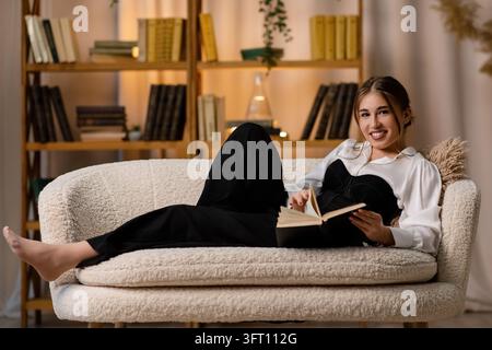 Lächelnde junge Frau liegt auf einem gemütlichen Sofa, liest ein Buch in einem stilvollen Wohnzimmer mit warmer Beleuchtung und modernen Regalen im Hintergrund. Stockfoto