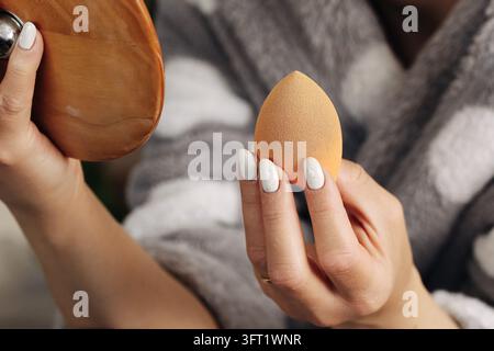Frau hält Make-up-Schwamm und Kompaktspiegel während der Schönheitsroutine. Kosmetik-Hintergrund für Grundierung, Gesichts-Make-up und persönliche Pflege Stockfoto