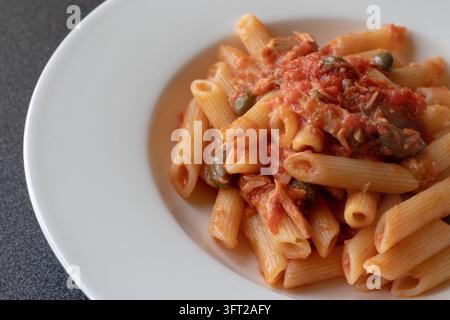 Hochwinkelansicht von Penne mit Tomatensoße, Thunfisch und Kapern in einer weißen Schüssel auf einer dunklen Arbeitsplatte, mit Thunfisch und Kapern obendrauf, weiches natürliches Licht Stockfoto