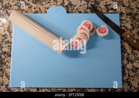 Hochwinkel-Ansicht von japanischem Narutomaki-Fischkuchen, der in spiralförmige Stücke geschnitten ist, neben einem Wellmessermesser auf einem blauen Plastikschnellbrett, Vorbereitung von Zutaten Stockfoto