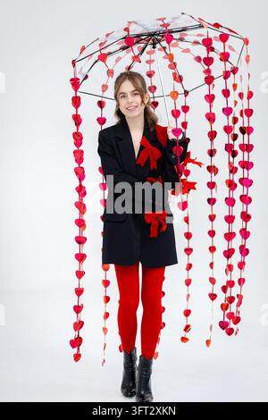 Lächelnde junge Frau hält einen Regenschirm mit roten herzförmigen Verzierungen, trägt einen schwarzen Blazer mit roten Schleifen und leuchtend rote Strumpfhosen. Stockfoto