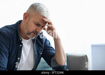 Frustrierter älterer bärtiger Mann sitzt auf dem Sofa im Wohnzimmer zu Hause, berührt seinen Kopf, hat finanzielle Probleme oder leidet unter Kopfschmerzen, Nahaufnahme Stockfoto