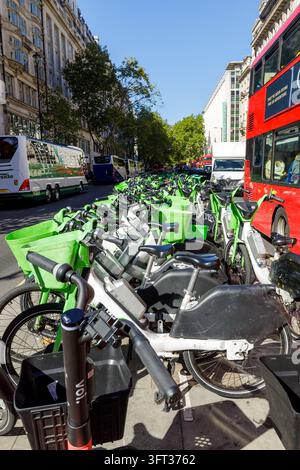 London, Vereinigtes Königreich - 21. September 2026: Blick auf eine Reihe geparkter limettengrüner Mietfahrräder und Elektroscooter auf einer belebten Stadtstraße mit einem roten Stockfoto