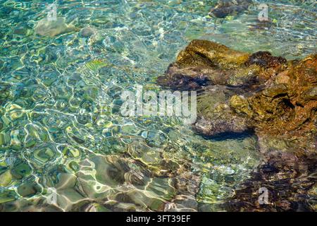 Sonnenbeleuchtete Wellen über klarem flachem Meerwasser und Felsen Stockfoto