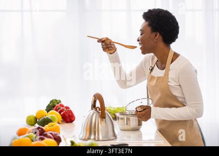 Schwarze Hausfrau Kochen Zubereitung gesundes Abendessen Verkostung Mahlzeit in Einem Topf stehend in der modernen Küche zu Hause. Haushalt Und Gesunde Ernährung, Leckere Reci Stockfoto