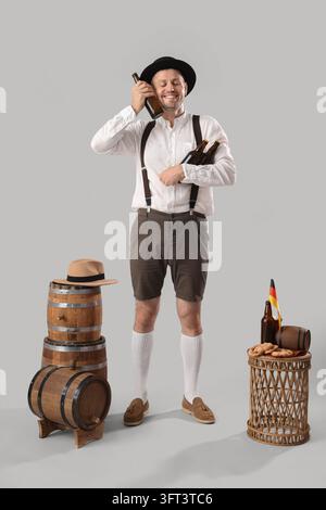 Oktoberfest-Feier und Bierkultur, Mann in traditioneller bayerischer Kleidung hält Glasflaschen mit Holzfässern und Brezeln auf grauem Hintergrund Stockfoto