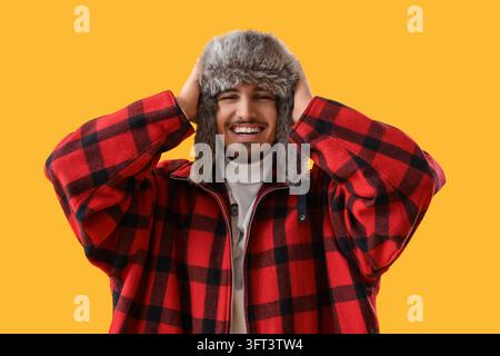 Wintermode und saisonale Freude, glücklicher junger Mann trägt eine rote karierte Jacke und einen Pelzmütze-Trapperhut und lacht auf einem leuchtend gelben Hintergrund Stockfoto