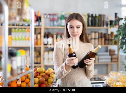 Nachdenkliche Einkäuferin betrachtet zwei Bierflaschen und vergleicht die Optionen beim Einkaufen von Getränken im örtlichen Supermarkt Stockfoto