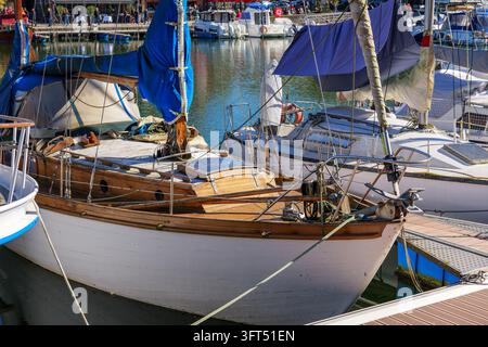 Holzsegelschiff in der Marina mit poliertem Deck, weißem Rumpf, blauer Segelabdeckung und umliegenden festgemachten Booten, die eine ruhige nautische Atmosphäre an einem stillen Tag schaffen Stockfoto