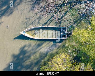 Einsames Boot ruht auf ruhigem, trübem Wasser. Äste werfen Schatten auf die Oberfläche. Grünes Laub rahmt den Rand der Szene ein Stockfoto