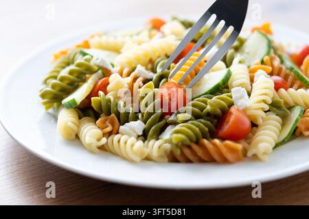 Nahaufnahme eines bunten Tricolor-Fusilli-Pasta-Salats mit Kirschtomaten, Zucchini und weißem Käse auf einem Teller. Gabel hält eine Kirschtomate in der Fo Stockfoto