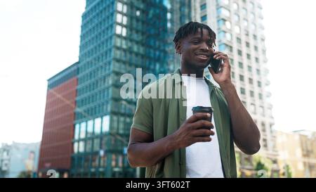 Junger Mann spricht am Smartphone, während er einen Takeaway-Kaffeebecher in einer modernen Stadtmitte hält Stockfoto
