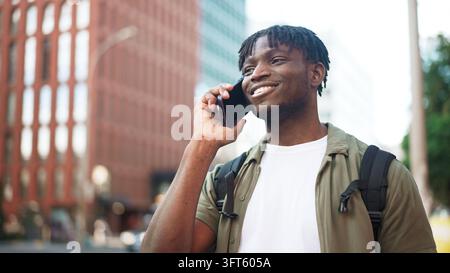 Lächelnder junger Mann mit Dreadlocks, der in der Innenstadt der Stadt auf einem Smartphone telefoniert, während er geht Stockfoto