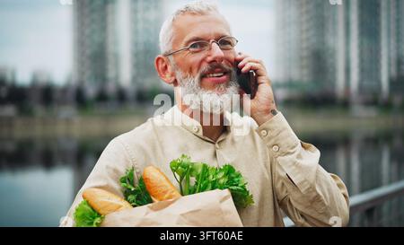 Reifer Mann mit grauem Bart, der an einem Smartphone spricht, während er eine Tasche mit frischen Lebensmitteln trägt Stockfoto