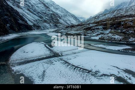 Luftaufnahme der Hussaini-Hängebrücke, die den eisigen Hunza-Fluss überspannt, umgeben von schneebedeckten Bergen und felsigen Ufern unter einem bewölkten Himmel in Gil Stockfoto
