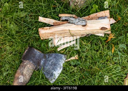 Gestapeltes gespaltenes Feuerholz und eine antike Axt, die zusammen auf grünem Gras in einem Hof ruhen. Eine Hochwinkelaufnahme zeigt gehackte Holzblöcke und den Kopf eines Metallbeils Stockfoto