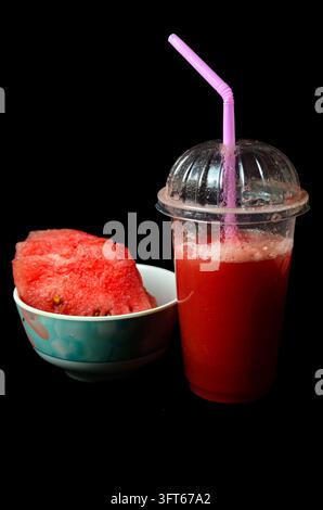 Frischer roter Wassermelonen-Smoothie in einem To-go-Becher mit einem rosa Strohhalm neben frischen Wassermelonenstücken in einer Schüssel, isoliert auf schwarzem Hintergrund. Stockfoto