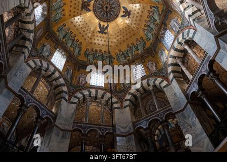 INTRIKATER GOLDENES MOSAIKKUPPEL, AACHENER DOM, AACHEN, DEUTSCHLAND: Ein direkter Blick nach oben auf das atemberaubend detaillierte goldene Mosaik an der oktogonalen Kuppeldecke der historischen alten Kathedrale. INFO: Der Aachener Dom, Deutschlands erstes UNESCO-Weltkulturerbe, ist ein karolingisches architektonisches Meisterwerk, das von Karl dem Großen gegründet wurde. Diese historische Sehenswürdigkeit, die byzantinische und gotische Elemente vereint, diente als Krönungskirche für einunddreißig deutsche Monarchen. Stockfoto