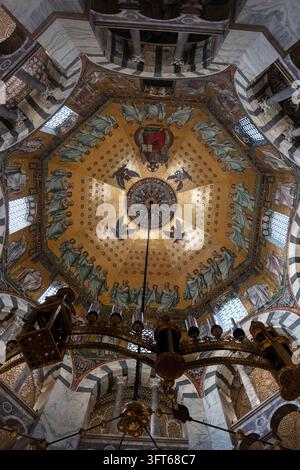 INTRIKATE GOLDENE MOSAIKKUPPEL UND KRONLEUCHTER, AACHENER DOM, AACHEN, DEUTSCHLAND: Ein direkter Blick nach oben auf das atemberaubend detaillierte goldene Mosaik an der achteckigen Kuppeldecke einer historischen Kapelle, mit einem massiven, kunstvollen Kronleuchter, der zentral hängt. INFO: Der Aachener Dom, Deutschlands erstes UNESCO-Weltkulturerbe, ist ein karolingisches Architekturmeisterwerk, das von Karl dem Großen gegründet wurde. Diese historische Stätte, die byzantinische und gotische Elemente vereint, diente als Krönungskirche für einunddreißig deutsche Monarchen. Stockfoto