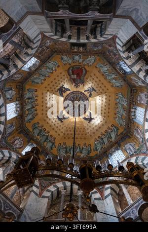 INTRIKATE GOLDENE MOSAIKKUPPEL UND KRONLEUCHTER, AACHENER DOM, AACHEN, DEUTSCHLAND: Ein direkter Blick nach oben auf das atemberaubend detaillierte goldene Mosaik an der achteckigen Kuppeldecke einer historischen Kapelle, mit einem massiven, kunstvollen Kronleuchter, der zentral hängt. INFO: Der Aachener Dom, Deutschlands erstes UNESCO-Weltkulturerbe, ist ein karolingisches Architekturmeisterwerk, das von Karl dem Großen gegründet wurde. Diese historische Stätte, die byzantinische und gotische Elemente vereint, diente als Krönungskirche für einunddreißig deutsche Monarchen. Stockfoto