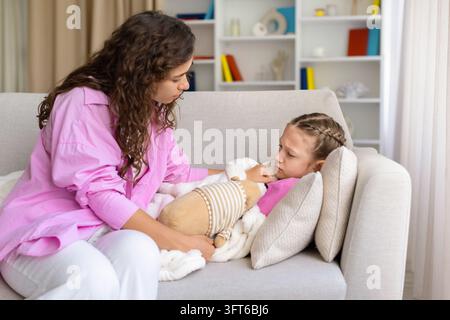 Försorgende Mutter tröstet ihre kranke Tochter, während das Mädchen unter einer warmen Decke mit einem weichen Spielzeug auf dem Sofa zu Hause liegt, und zeigt Unterstützung und Zärtlichkeit während Stockfoto