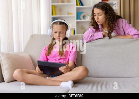 Mutter beobachtet ihre Tochter, die mit Kopfhörern ein Tablet benutzt, während sie zu Hause sitzt Stockfoto