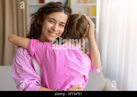 Lächelnde Mutter umarmt ihre Tochter, während sie zu Hause auf dem Sofa sitzt Stockfoto