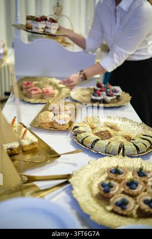 Eine Person in einem weißen Hemd ordnet Tabletts mit Kuchen, Gebäck und Obstkuchen auf einem Buffet-Tisch bei einem Ball an. Stockfoto