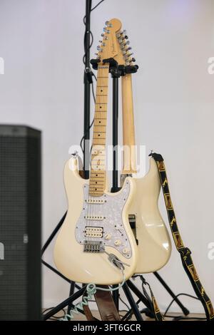 Eine cremefarbene E-Gitarre ruht auf einem Ständer vor einer weiteren Gitarre neben Bühnenausrüstung bei einem Ball. Stockfoto