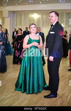 Eine Frau in einem langen grünen Kleid steht neben einem Mann in einem dunklen Anzug und einer Brille zwischen Gästen auf der Tanzfläche bei einem Ball. Stockfoto