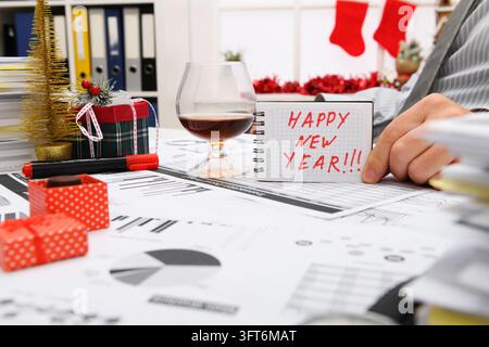 Geschäfts- und Urlaubskonzept – Geschäftsmann feiert in einem Büro, das mit Weihnachtsbaum, Süßigkeiten und anderen Neujahrsdekorationen geschmückt ist. Zeigt ein Notizbuch Stockfoto