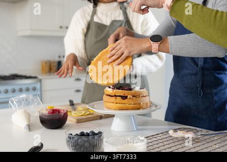 Verschiedene Freundinnen legen die obere Biskuitsschicht auf einen weißen Kuchenständer in der Küche und tragen Schürzen Stockfoto