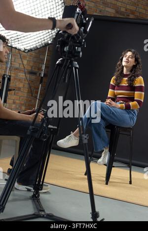 Kleine Crew filmt sitzende Frau mit gestreiftem Pullover, Jeans auf Hocker nahe Kamerastativ, Softbox. Studio, Interview, Moderatorin, Beleuchtung, Hintergrund, Stockfoto
